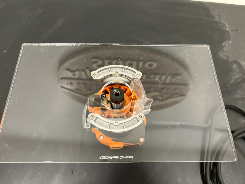 Peut inclure: Un gros plan sur une fraise de toupie fix&eacute;e &agrave; une base de toupie en acrylique transparente. La base porte l'inscription "Compact Router Base".