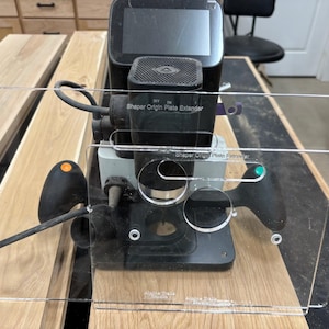 Peut inclure: Un prolongateur de plaque Shaper Origin noir avec une plaque en acrylique transparente, monté sur une surface en bois. Le prolongateur a un écran numérique et des boutons de commande. Le texte "Shaper Origin Plate Extender" est visible.