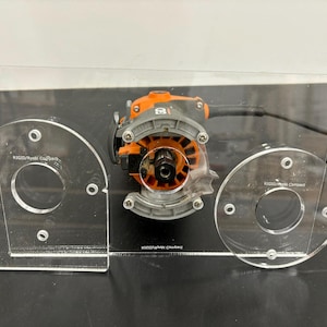 May include: A clear acrylic router template with two circular openings and the text "Kutzall Wood Company" printed on each opening. A black and orange router is centred in the template.