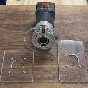 May include: A dark blue and silver handheld power router with a circular clear acrylic base plate attached, accompanied by two additional clear acrylic base plates, one square and one rectangular, each featuring four pre-drilled mounting holes.