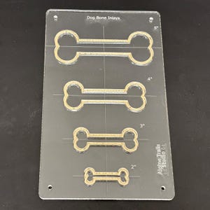 K&ouml;nnte beinhalten: Eine transparente Kunststoff-Schablone mit vier Hundebeinknochenformen in verschiedenen Gr&ouml;&szlig;en. Die Schablone ist mit Ma&szlig;en in Zoll gekennzeichnet: 2", 3", 4" und 5". Der Text "Dog Bone Inlays" und "Alpine Trails Studio LA" ist auf der Schablone aufgedruckt.