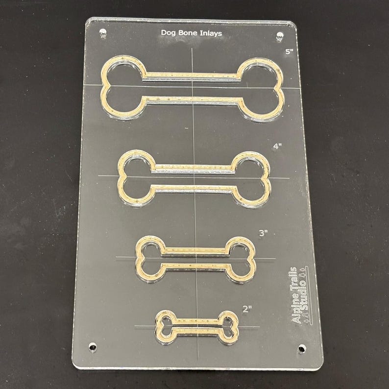 K&ouml;nnte beinhalten: Eine transparente Kunststoff-Schablone mit vier Hundebeinknochenformen in verschiedenen Gr&ouml;&szlig;en. Die Schablone ist mit Ma&szlig;en in Zoll gekennzeichnet: 2", 3", 4" und 5". Der Text "Dog Bone Inlays" und "Alpine Trails Studio LA" ist auf der Schablone aufgedruckt.