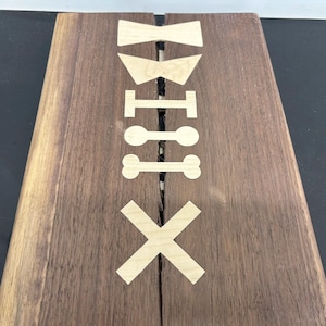 K&ouml;nnte beinhalten: Ein Holzbrett mit einem dekorativen Design, das aus dem Holz geschnitten ist. Das Design umfasst eine Fliege, einen Kragen, einen Knochen und ein X. Das Brett ist in der Mitte gespalten.