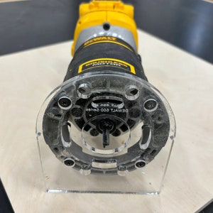 May include: A black and yellow DeWalt 600 series router with a clear plastic base and a clear plastic template guide. The router is on a light brown surface.