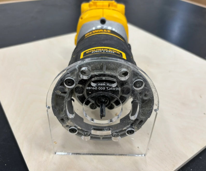 May include: A black and yellow DeWalt 600 series router with a clear plastic base and a clear plastic template guide. The router is on a light brown surface.