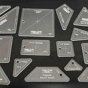 May include: A set of 12 clear acrylic quilting rulers with various shapes and sizes, including squares, rectangles, triangles, and a trapezoid. The rulers are marked with measurements in inches and have the text "Alpine Trails Studio" printed on them.