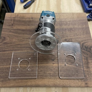 Könnte beinhalten: Eine türkisfarbene und silberne Makita Kompaktfräse mit montierter runder, transparenter Acryl-Grundplatte, begleitet von zwei weiteren transparenten Acryl-Grundplatten, einer quadratischen und einer rechteckigen, jeweils mit zentraler Öffnung.