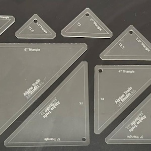 May include: A set of nine clear plastic triangle rulers for quilting. The rulers are labelled with their size in inches and have a hole at the top. The rulers are arranged in a grid pattern on a black surface.