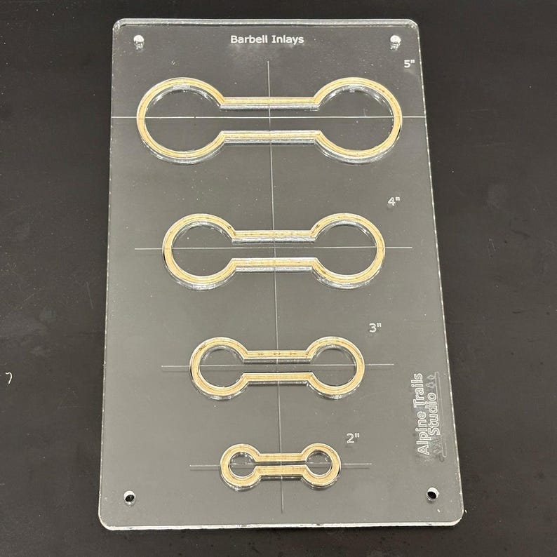 K&ouml;nnte beinhalten: Eine transparente Kunststoffschablone mit vier verschiedenen Gr&ouml;&szlig;en von Barbell-Einlegemustern. Die Schablone ist mit "Barbell Inlays" beschriftet und enth&auml;lt Ma&szlig;e in Zoll: 5", 4", 3" und 2". Die Schablone ist auch mit "Alpine Trails Studio 52" beschriftet.