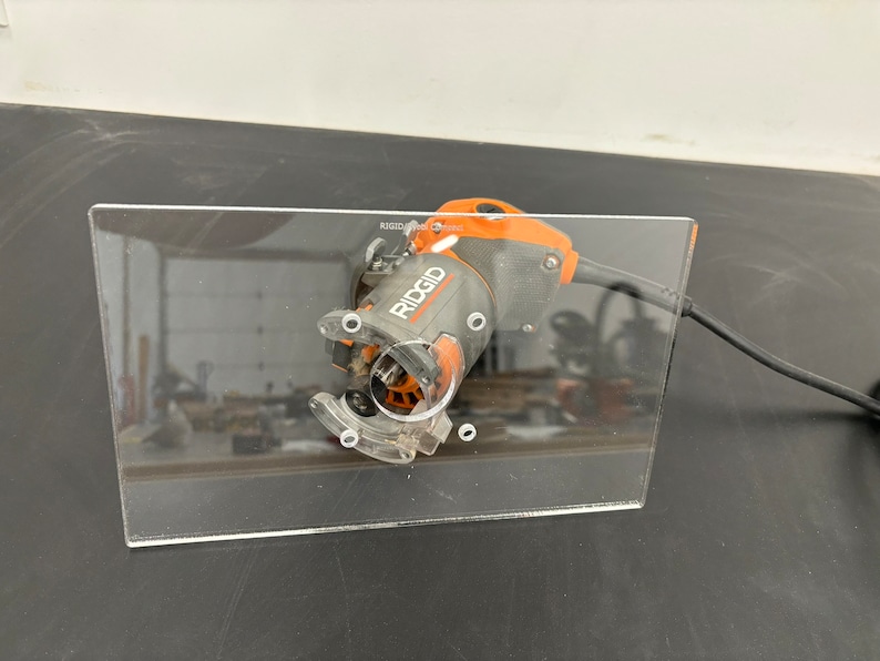 Peut inclure: Une base de d&eacute;fonceuse en acrylique transparente avec une d&eacute;fonceuse Ridgid noire et orange fix&eacute;e. La d&eacute;fonceuse est branch&eacute;e et le cordon est visible.