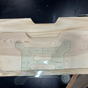 Peut inclure: Un gabarit en plastique transparent pour projets de menuiserie est placé sur une surface en bois clair. Le gabarit a une forme découpée avec plusieurs trous et fentes.