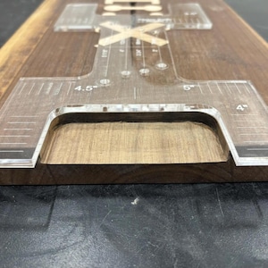 May include: A clear acrylic woodworking jig with measurement markings in inches, including 4", 4.5", and 5". The jig is positioned on a dark wood surface, with a routed-out section visible. The tool is designed for precise woodworking tasks.
