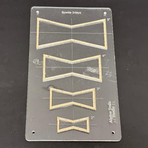 K&ouml;nnte beinhalten: Eine transparente Kunststoff-Schablone mit vier Fliegenbindeformen f&uuml;r Einlegearbeiten. Die Schablone ist mit "Bowtie Inlays" beschriftet und enth&auml;lt Ma&szlig;e in Zoll: 5", 4", 3" und 2". Die Schablone ist auch mit "Alpine Trails Studio 21" beschriftet.