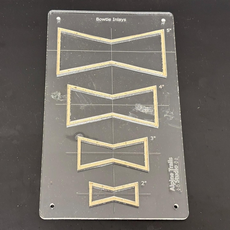K&ouml;nnte beinhalten: Eine transparente Kunststoff-Schablone mit vier Fliegenbindeformen f&uuml;r Einlegearbeiten. Die Schablone ist mit "Bowtie Inlays" beschriftet und enth&auml;lt Ma&szlig;e in Zoll: 5", 4", 3" und 2". Die Schablone ist auch mit "Alpine Trails Studio 21" beschriftet.
