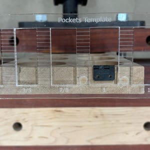May include: A clear plastic template with the words "Pockets Template" printed on it. The template has markings for 1/2 inch, 5/8 inch, 3/5 inch, 7/8 inch, 1 inch, 1 1/8 inch, and 1 1/4 inch. The template is being used to create pockets in a piece of wood.