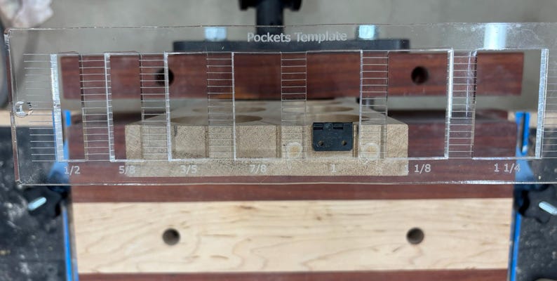 May include: A clear plastic template with the words "Pockets Template" printed on it. The template has markings for 1/2 inch, 5/8 inch, 3/5 inch, 7/8 inch, 1 inch, 1 1/8 inch, and 1 1/4 inch. The template is being used to create pockets in a piece of wood.