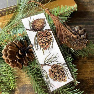 May include: A glass bookmark with a brown tassel. The bookmark features a white background with three hand-painted pine cones on branches.