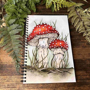 May include: A spiral-bound notebook featuring a watercolor illustration of two red and white spotted mushrooms. The mushrooms have thick, light-colored stems and are surrounded by thin, green, grass-like blades on a light, neutral background.