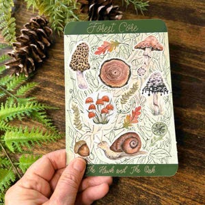 May include: A green card with the text "Forest Core" and "The Hawk and The Oak". The card features illustrations of mushrooms, leaves, a snail, and an acorn. The card is held in a person's hand.