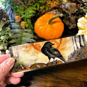 May include: A rectangular bookmark featuring a watercolor illustration of a black crow perched on pumpkins against a textured orange and grey background. The artwork includes a large orange moon and the text Hawk and the Oak on the right side.