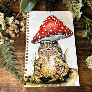 May include: A spiral-bound notebook featuring a watercolor illustration of a textured brown toad sitting beneath a large red mushroom with white spots. The toad has detailed skin markings and is positioned in a grassy area on a white background.