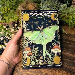 May include: A spiral-bound notebook with a dark blue cover featuring a large green moth illustration. The design includes mushrooms, moons, stars, and mountains. The notebook has a gold spiral binding.