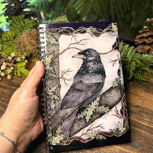 May include: A spiral-bound notebook featuring a detailed illustration of a crow perched on a tree branch. The artwork is framed with a border of leaves and branches, with a dark blue cover. The crow is depicted in shades of gray and black.