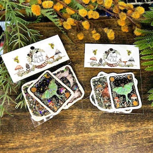 May include: Two sets of stickers with a nature theme. Each set includes stickers with a moth, mushrooms, and the text "Hawk & Oak". The stickers have a dark background with a gold border. The background is a wooden surface with yellow flowers and green leaves.