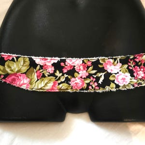 Peut inclure: Une ceinture en tissu noir avec un motif floral de roses roses et de feuilles vertes. La ceinture a un bord blanc effiloché. La ceinture est présentée sur un mannequin noir.