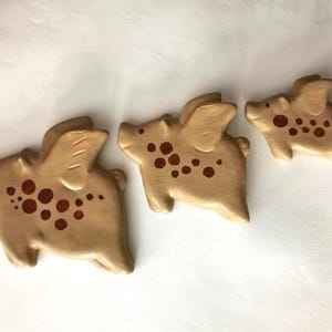 Puede incluir: Tres figuras caprichosas de cerdos de color beige con alas y manchas de color rojo oscuro. Los cerdos varían en tamaño, el más grande a la izquierda y el más pequeño a la derecha. Están dispuestos sobre una superficie blanca.
