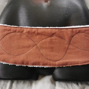 Peut inclure: Une ceinture en tissu matelassé marron avec une doublure en polaire blanche. La ceinture a un motif cousu ondulé.