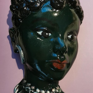 May include: A dark green ceramic sculpture of a person's head with a detailed hairstyle and a silver necklace. The sculpture has a realistic face with painted eyes, nose, and lips.