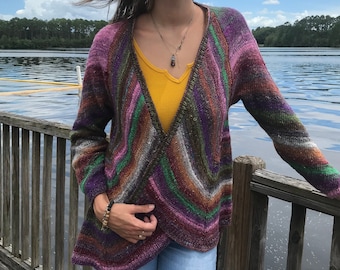 May include: A woman wearing a multi-coloured, long-sleeved, knitted cardigan with a deep V-neck. The cardigan has a striped pattern with shades of purple, green, brown, and pink. She is wearing a yellow vest top underneath and blue jeans.