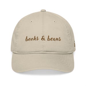 May include: A beige baseball cap with the words "books & beans" embroidered in brown script across the front. The cap has a curved brim and a structured crown. The hat is a casual accessory.