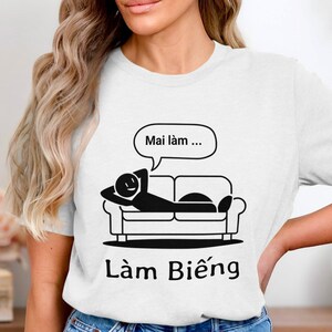 May include: White t-shirt with a black graphic of a stick figure lounging on a couch, with a speech bubble above saying "Mai làm..." and the word "Làm Biếng" below. The design is simple and bold.