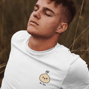May include: White t-shirt featuring a cartoon dumpling with a smiling face and the text "Bao Wow" in a handwritten style. The dumpling has steam lines above it. The shirt is made of a soft material.