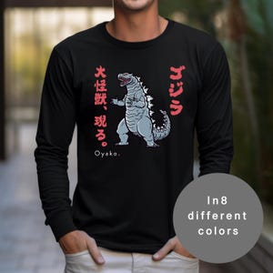 May include: Black long-sleeve shirt with a Godzilla illustration in grey and white, and red Japanese text. The shirt also features the text "Oyako" and a circular graphic that says "In 8 different colours."