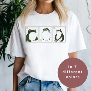 Könnte beinhalten: Weißes T-Shirt mit einem Aufdruck von drei Fröschen in schwarzer Tinte. Die Frösche sind in verschiedenen Posen, einer sitzt, einer steht und einer schaut nach oben. Der Text "In 7 verschiedenen Farben" befindet sich auf einem braunen Kreis.