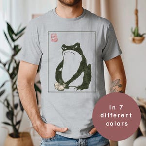 May include: A light gray t-shirt featuring a frog illustration in a Japanese art style. The frog is dark green and holds a small bowl. The design is framed in a black rectangle with a red stamp in the upper left corner. The shirt is available in 7 different colors.