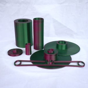 Può includere: Un set di parti in plastica stampate in 3D di colore verde e rosa per una stampante 3D. Il set comprende due cilindri, un piccolo cilindro, un disco rotondo, un disco più grande con un disco più piccolo sopra e un pezzo lungo, sottile e curvo.