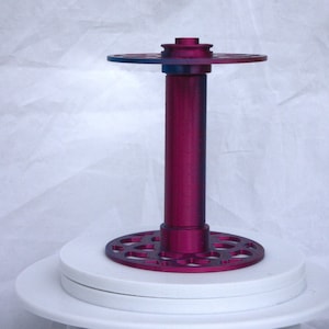 Può includere: Un porta bobina metallico color magenta con una colonna centrale cilindrica e dischi traforati circolari in alto e in basso. Il supporto è posto su una piattaforma bianca a strati, con uno sfondo bianco morbido.