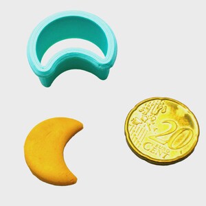 May include: A teal plastic crescent moon cookie cutter next to a baked crescent moon cookie and a 20 Euro cent coin. The cookie cutter is made of multiple layers of plastic. The coin is gold-coloured.