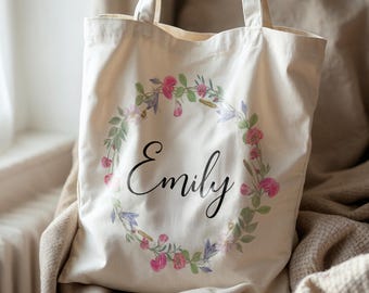 May include: Cream-colored tote bag with a floral wreath design and the name "Emily" in elegant script. The wreath features pink and purple flowers with green leaves. The bag has two handles.
