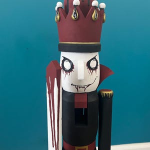 Pode incluir: Um quebra-nozes de madeira pintado à mão com tema de vampiro. O quebra-nozes tem um rosto branco com maquiagem preta nos olhos e um sorriso sinistro. Ele usa uma coroa bordô e uma roupa preta com cinto vermelho. Ele segura uma espada vermelha e branca.