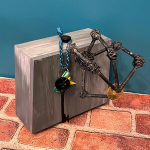 May include: A gray rectangular block with a black key hanging from a blue cord. A metal sculpture with a yellow disc is attached to the block. The background is blue and the surface is brick.
