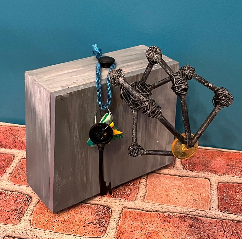 May include: A gray rectangular block with a black key hanging from a blue cord. A metal sculpture with a yellow disc is attached to the block. The background is blue and the surface is brick.