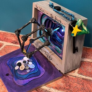 May include: A mixed-media art piece featuring a wooden box with the words "Be Wise, Be Brave, Be Tricky." A key with a star and a green triangle hangs from the box. A sculpture with buttons and wire extends from a purple base.