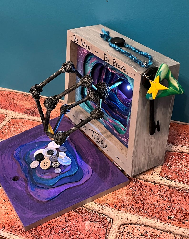 May include: A mixed-media art piece featuring a wooden box with the words "Be Wise, Be Brave, Be Tricky." A key with a star and a green triangle hangs from the box. A sculpture with buttons and wire extends from a purple base.