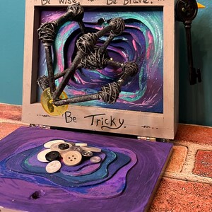 May include: A mixed-media art piece with a wooden frame, featuring the phrases "Be Wise, Be Brave, Be Tricky." Inside, wire-wrapped nails and a key are set against a layered, swirling background of purple, teal, and green. The lower section has layered purple with buttons.
