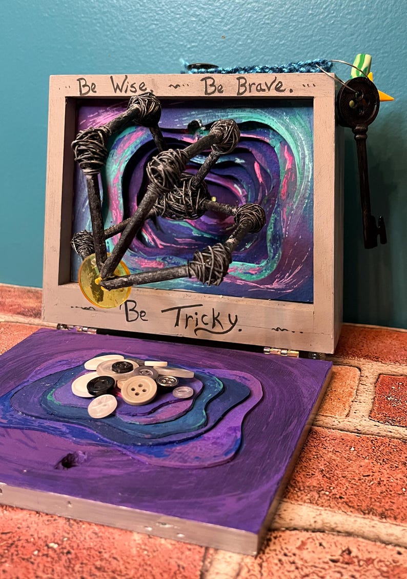 May include: A mixed-media art piece with a wooden frame, featuring the phrases "Be Wise, Be Brave, Be Tricky." Inside, wire-wrapped nails and a key are set against a layered, swirling background of purple, teal, and green. The lower section has layered purple with buttons.